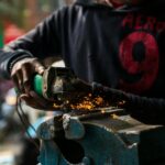 Crop anonymous African American male worker in dirty wear welding metal details in blurred factory
