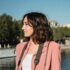 Profile of a woman in a pink blazer by a riverside on a sunny day.